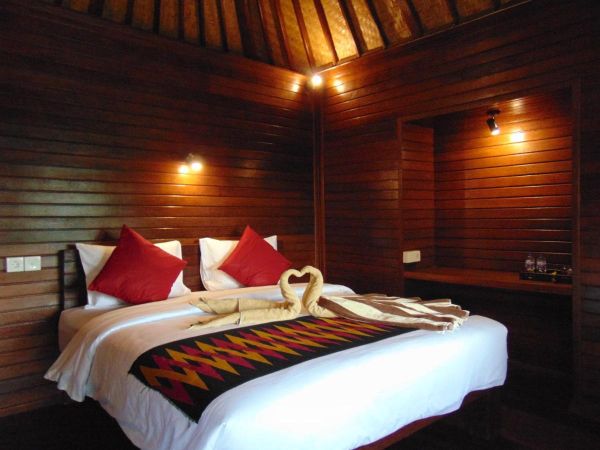 Cozy Balinese bungalow bedroom with wooden walls, thatched ceiling, king bed featuring swan towel art, red pillows, and warm ambient lighting. - Bali Villas